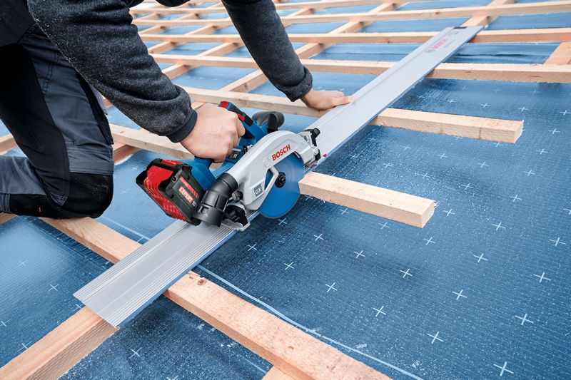 A person using a circular saw to cut a metal sheet on a rooftop frame.