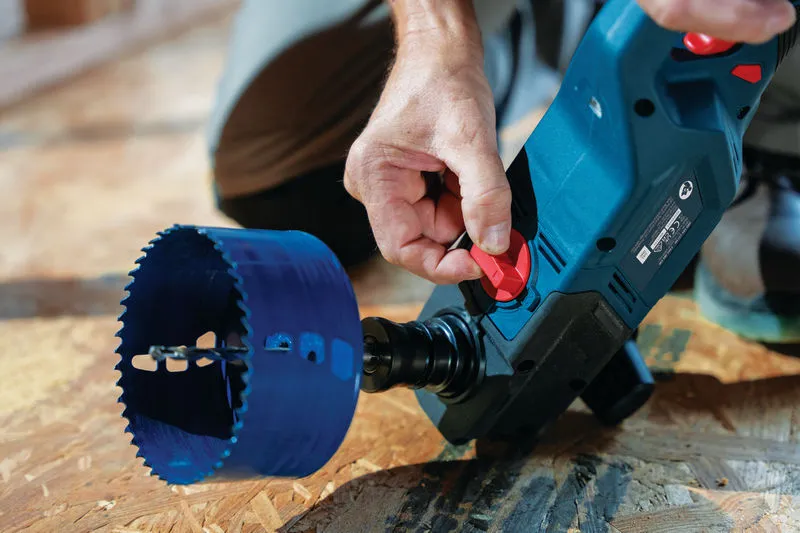 A person adjusts a cordless angle drill with a large hole saw on a wooden surface.