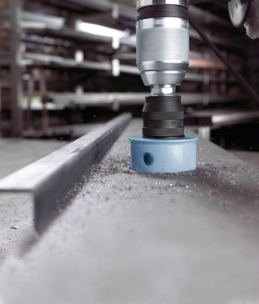 Hole saw attachment drilling through a steel beam, metal shavings scattered on the surface.