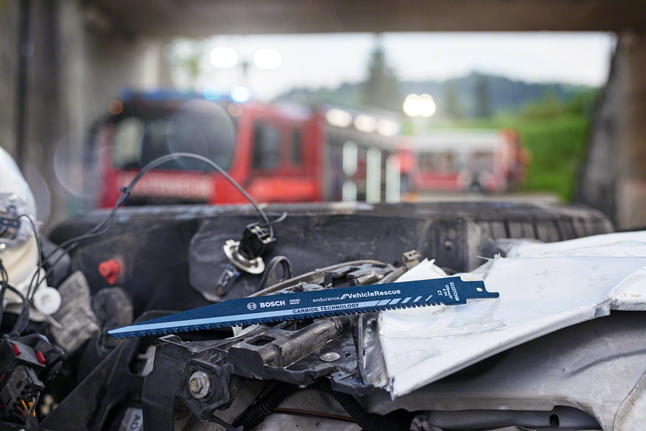 Rescue saw blade lying on damaged car hood at accident scene with emergency vehicles.