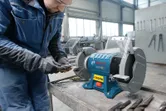 A person wearing safety equipment sharpens metal using a double-wheeled bench grinder.