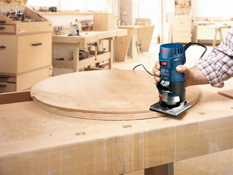 Person guides a palm router along the edge of a round wooden board on a workbench.