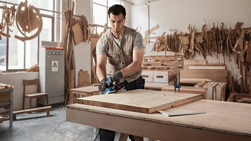 A person wearing safety equipment planes a wooden board in a workshop.