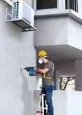 A person wearing safety equipment drills into a wall beneath an air conditioning unit.