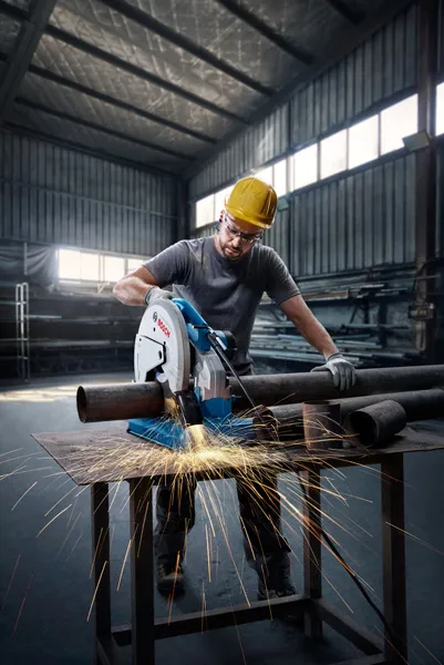 A person wearing safety equipment cuts steel pipes, creating sparks with a power saw.