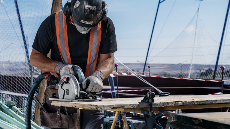 A person wearing safety equipment cuts wood with a circular saw at a construction site.