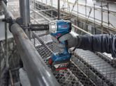 A person wearing safety equipment uses a cordless impact wrench on scaffolding.