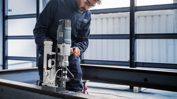A person wearing safety equipment operates a magnetic drill on a steel beam.
