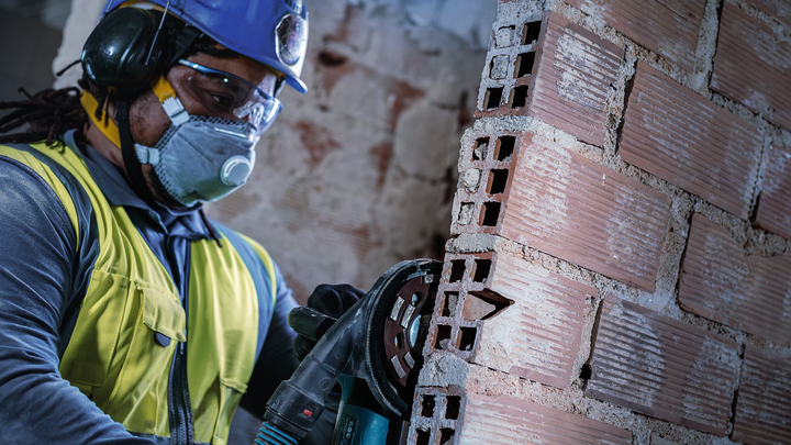 A person wearing safety equipment cuts a brick wall with a power tool.