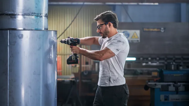 Person wearing safety equipment drills into a large metal cylinder in a workshop.
