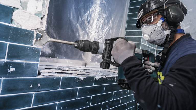 A person wearing safety equipment removes tiles with a demolition hammer.
