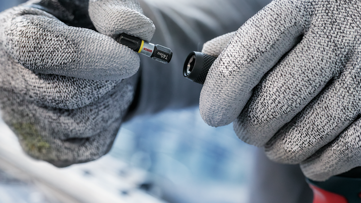 Person wearing safety equipment holds a screwdriver bit near a drill attachment.