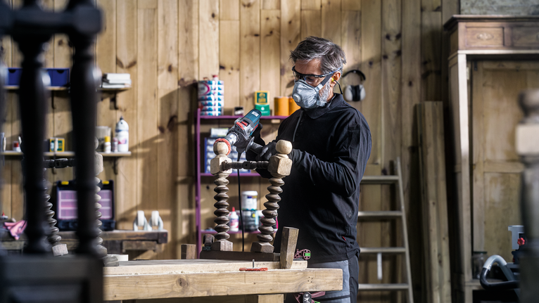 Person wearing safety equipment polishes wooden furniture in a workshop.