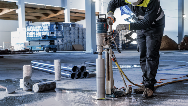 A person wearing safety equipment drills a hole into concrete on a construction site.