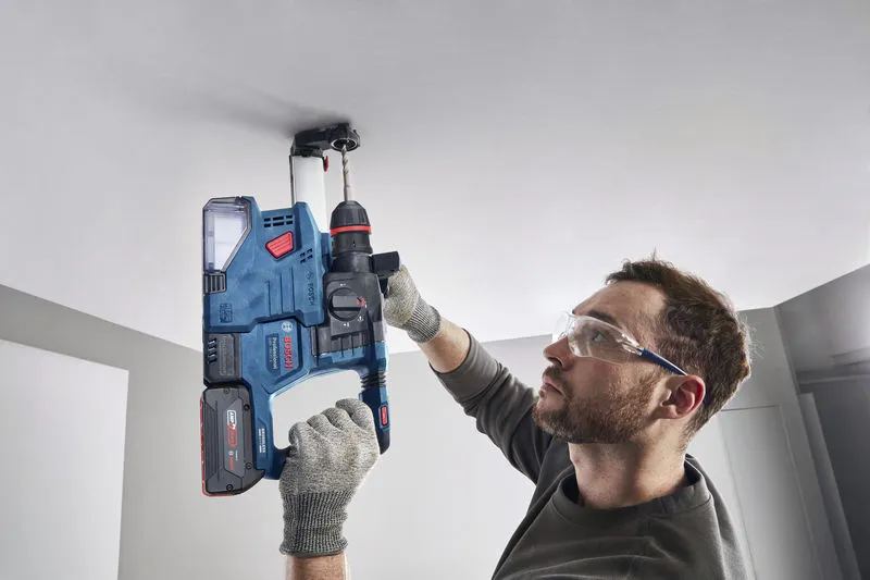 A person wearing safety equipment drills into a ceiling using a cordless rotary hammer.