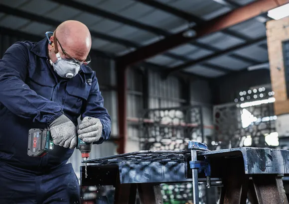 A person wearing safety equipment drills into a metal surface in a workshop.