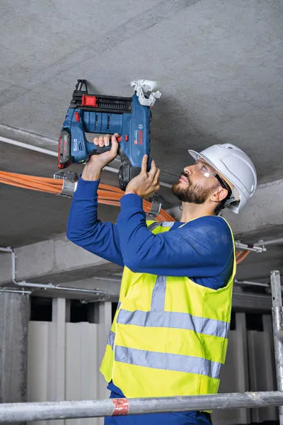 A person wearing safety equipment uses a cordless concrete nailer on a ceiling.