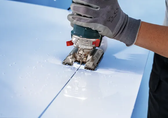 Worker wearing safety equipment uses a jigsaw to cut a straight line in a white panel.
