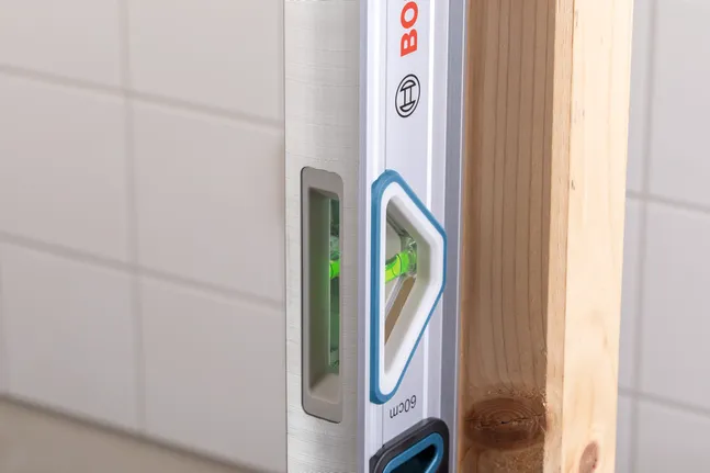 Spirit level checks vertical alignment of a wooden beam against a tiled wall.