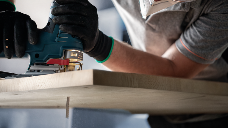 Person wearing safety equipment uses a jigsaw to cut a wooden board.