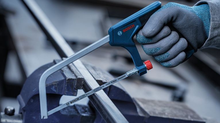 Person wearing safety equipment uses a hacksaw to cut a metal bar.