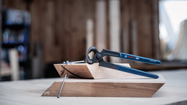 Pincers resting on wooden blocks beside a large nail on a workshop table.