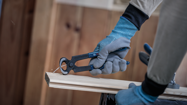 Person wearing safety equipment pulls a nail from a wooden board with pliers.