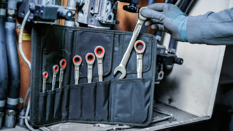 Person wearing safety equipment selects a wrench from a tool pouch near machinery.