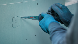 Person wearing safety equipment cuts a marked square in drywall with a hand saw.