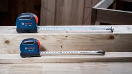 Two tape measures extended on stacked wooden beams in a workshop.