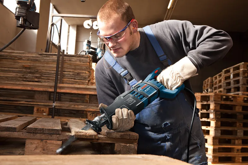 A person wearing safety equipment uses a reciprocating saw to cut a wooden pallet.