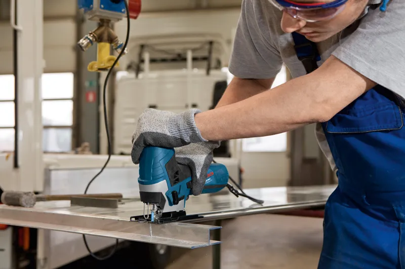 A person wearing safety equipment cuts a metal sheet using a jigsaw.