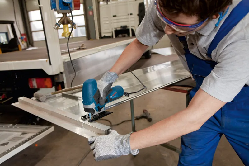 A person wearing safety equipment uses a jigsaw to cut a sheet of metal.