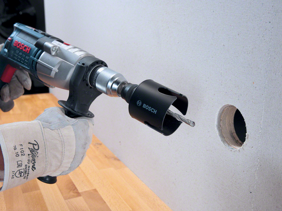 Worker wearing safety equipment drills a circular hole into drywall with a power tool.