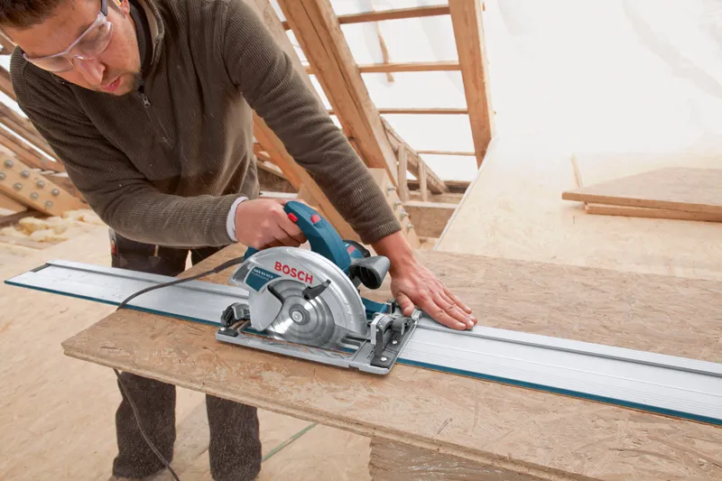 A person wearing safety equipment guides a circular saw along a rail to cut wood.