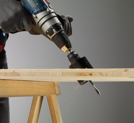 Une personne portant un équipement de sécurité utilise une perceuse électrique munie d'une scie cloche pour découper du bois.