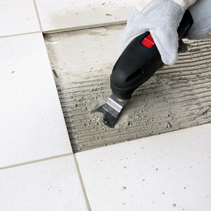 Un travailleur portant un équipement de sécurité utilise un outil multifonction pour retirer l'adhésif entre les carreaux de sol.