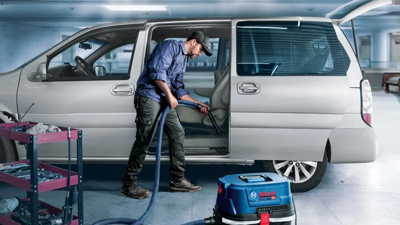 Une personne nettoie l'intérieur d'une voiture avec un aspirateur à côté d'un chariot à outils dans un garage.