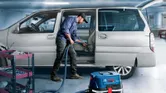 Une personne nettoie l'intérieur d'une voiture avec un aspirateur à côté d'un chariot à outils dans un garage.