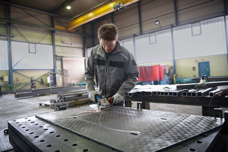 Une personne portant un équipement de sécurité broie une grande feuille de métal dans un espace de travail d'usine.
