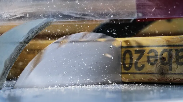 Une scie circulaire coupe une poutre en bois, de la sciure vole dans l'air.