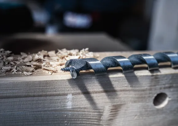 Grande mèche à tarière posée sur une planche de bois avec des copeaux de bois à côté.