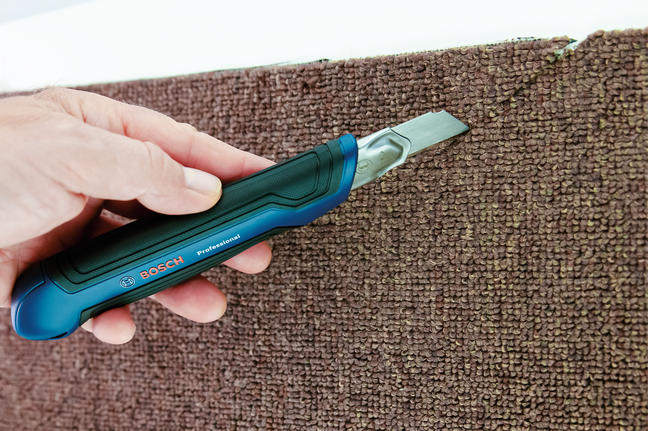 Une personne découpe un tapis marron le long du bord à l'aide d'un cutter.