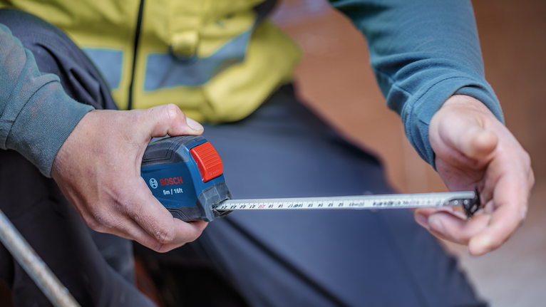 La personne portant un équipement de sécurité utilise un mètre ruban pour vérifier la distance.