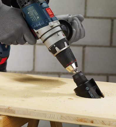 Person wearing safety equipment uses a power drill with a hole saw to cut wood.