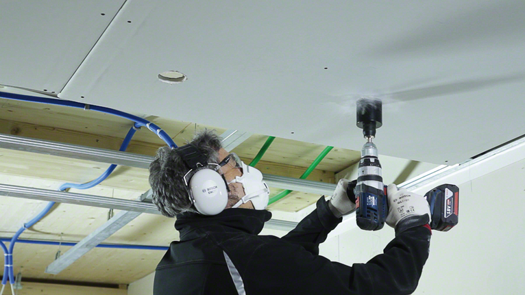 Person wearing safety equipment drills a hole in a ceiling panel.