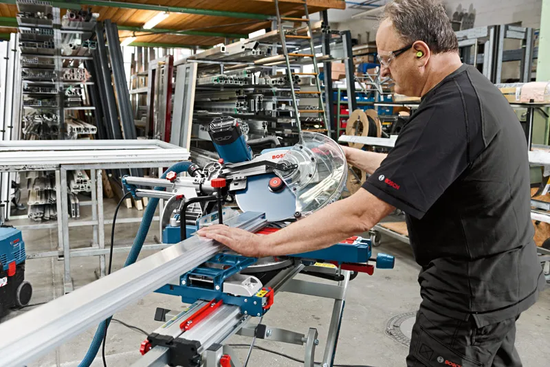 A person wearing safety equipment operates a sliding mitre saw to cut a metal profile.