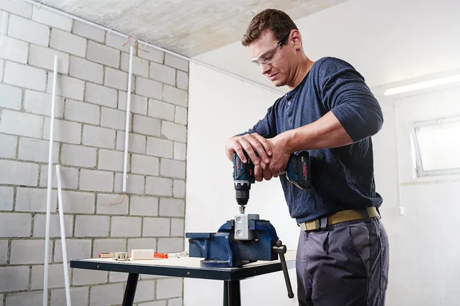 Person wearing safety equipment uses a power drill on metal clamped in a vice.