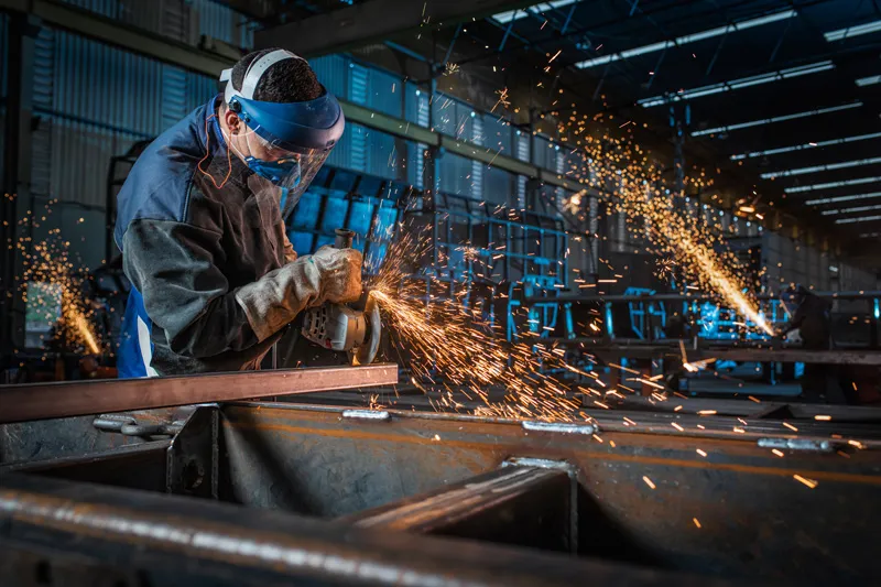 A person wearing safety equipment grinds metal, producing bright sparks in a factory.