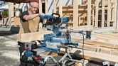 A person wearing safety equipment operates a sliding mitre saw to cut wooden beams.
